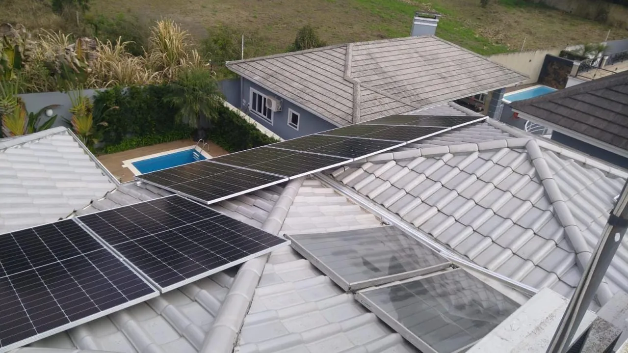 Módulos fotovoltaicos sobre telhado metálico de residência, com piscina e área verde ao fundo